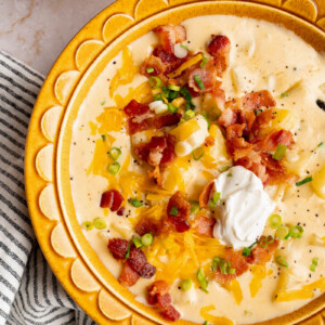 Loaded baked potato soup in a yellow bowl on a table with chopped bacon, shredded cheddar cheese, sliced green onions and soup cream on top.