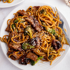Beef and brroccoli lo mein noodles on a white plate for serving. White sesame seeds are sprinkled on top.