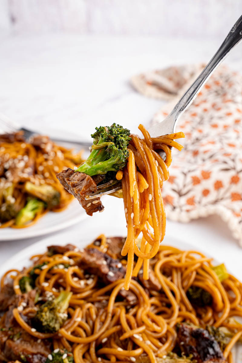 A fork lifts up noodles with a piece of broccoli and steak before eating.