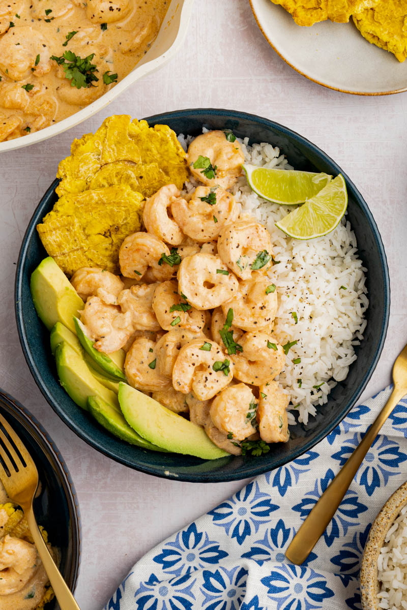 Camarones al ajillo in a bowl on top of white rice, with a side of tostones and sliced avocado.