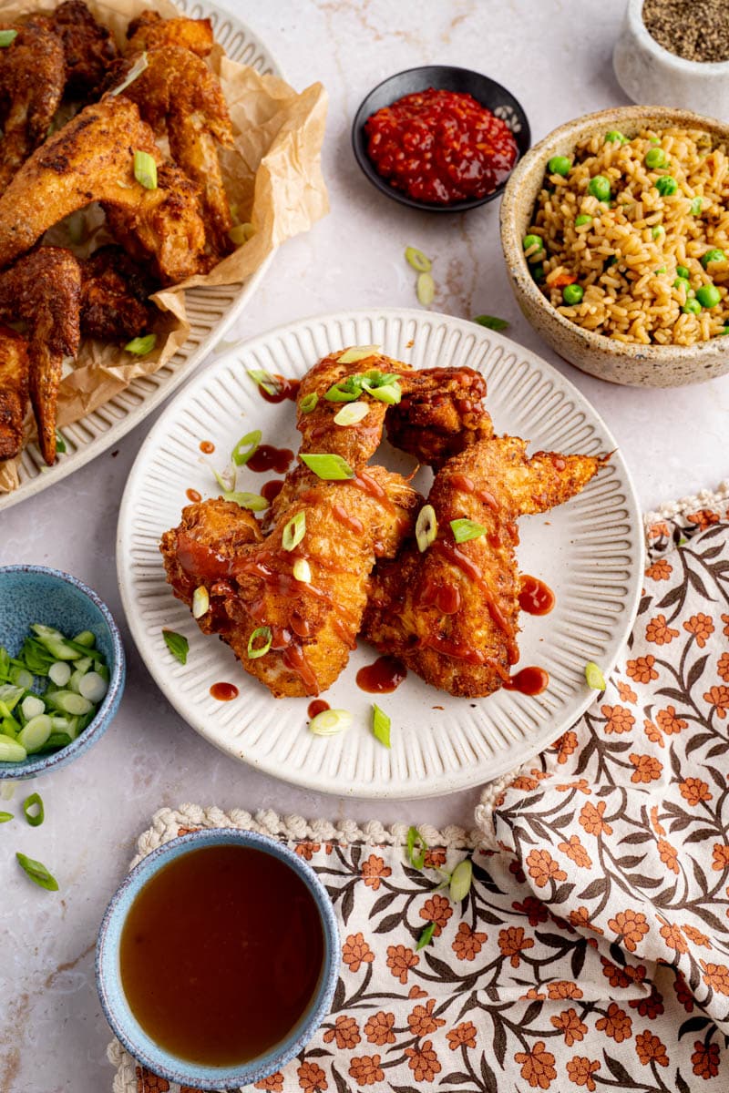 Three fried chicken wings on a plate with some hot sauce drizzled on top for serving. The remaining wings are on a plate to the side and a bowl of fried rice also sits next to the weeks on a table.