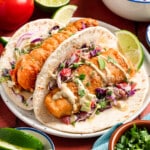 Two baja fish tacos on a white plate on a table with fresh slaw in a bowl to the side.