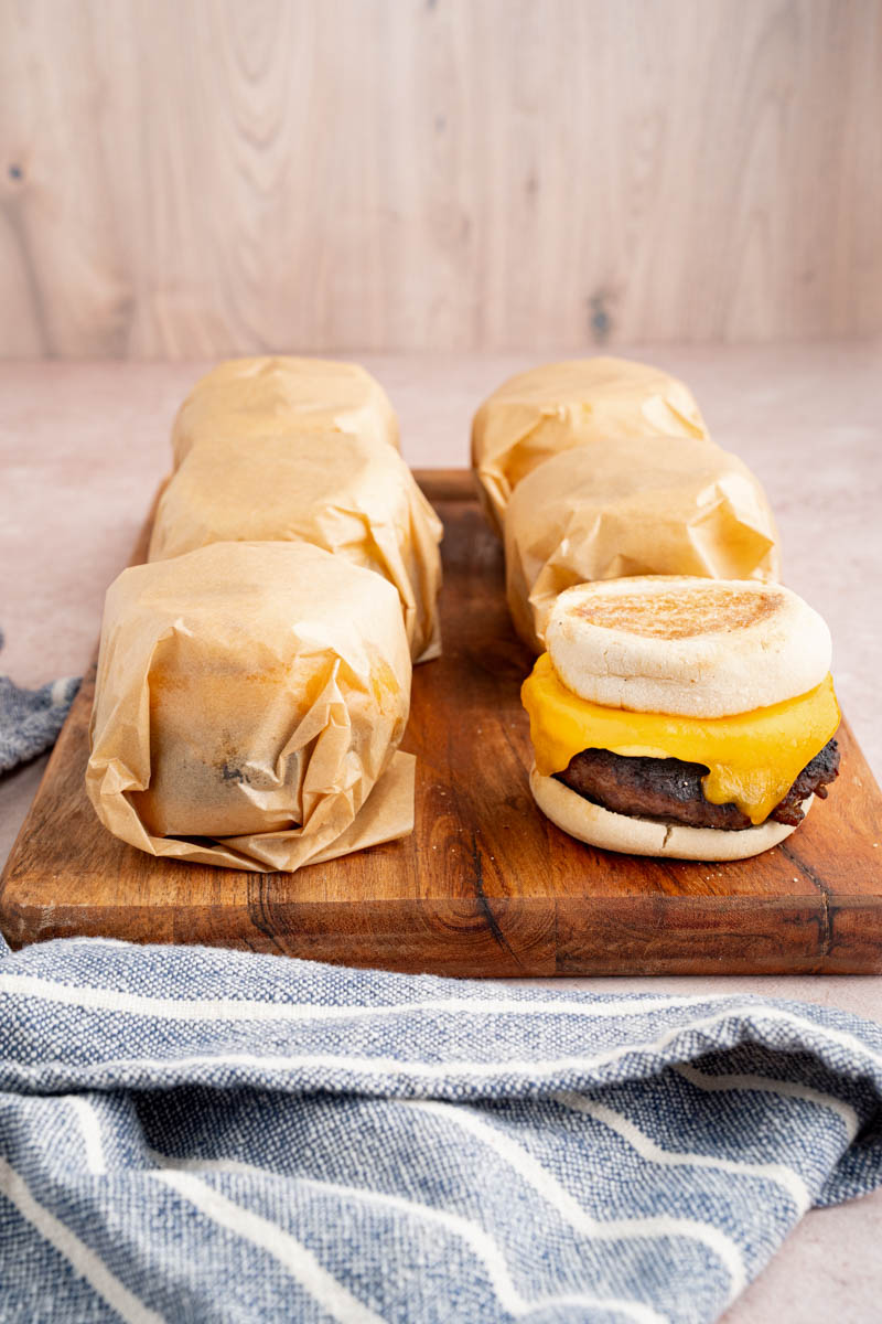 Five breakfast sandwiches that are wrapped in parchment paper sit on a wooden cutting board.