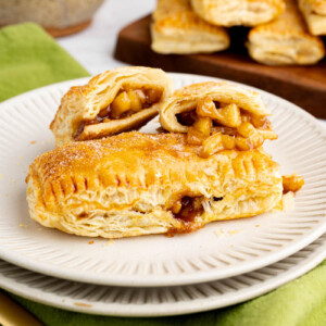 An apple hand pie split into two pieces sits next to a whole apple hand pie on a plate for serving.