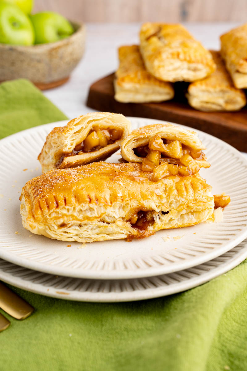 An apple hand pie split into two pieces sits next to a whole apple hand pie on a plate for serving.