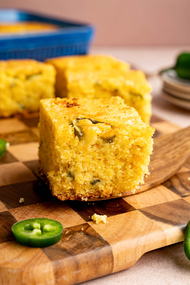 A serving utensil holds a piece of jalapeno cheddar cornbread on top of a wooden cutting board for serving.