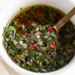 A small white bowl filled with chimichurri sauce. A spoon is scooping out some of the sauce from the bowl.