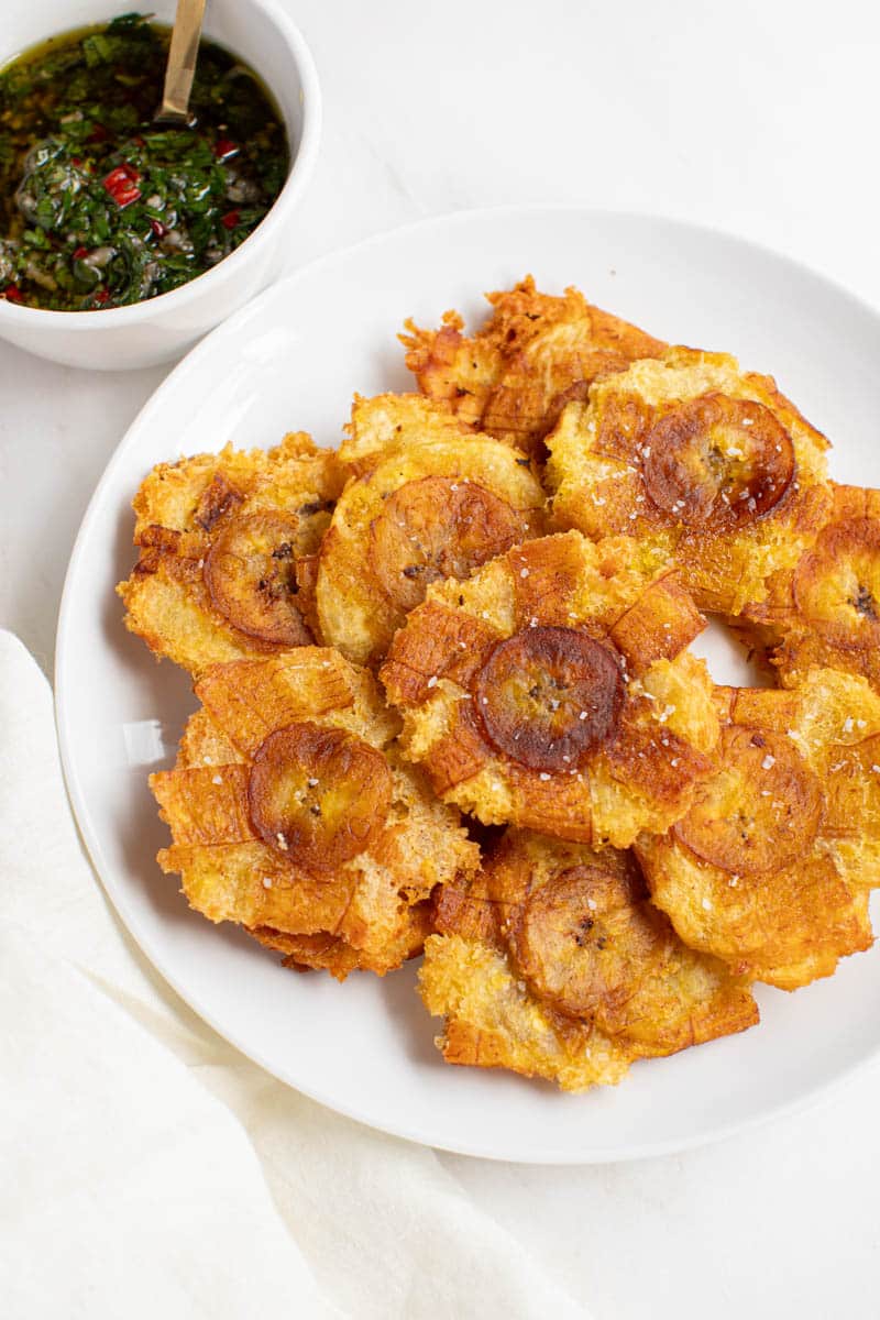 Fried green plantains (tostones) on a plate next to a bowl of chimichurri for serving.