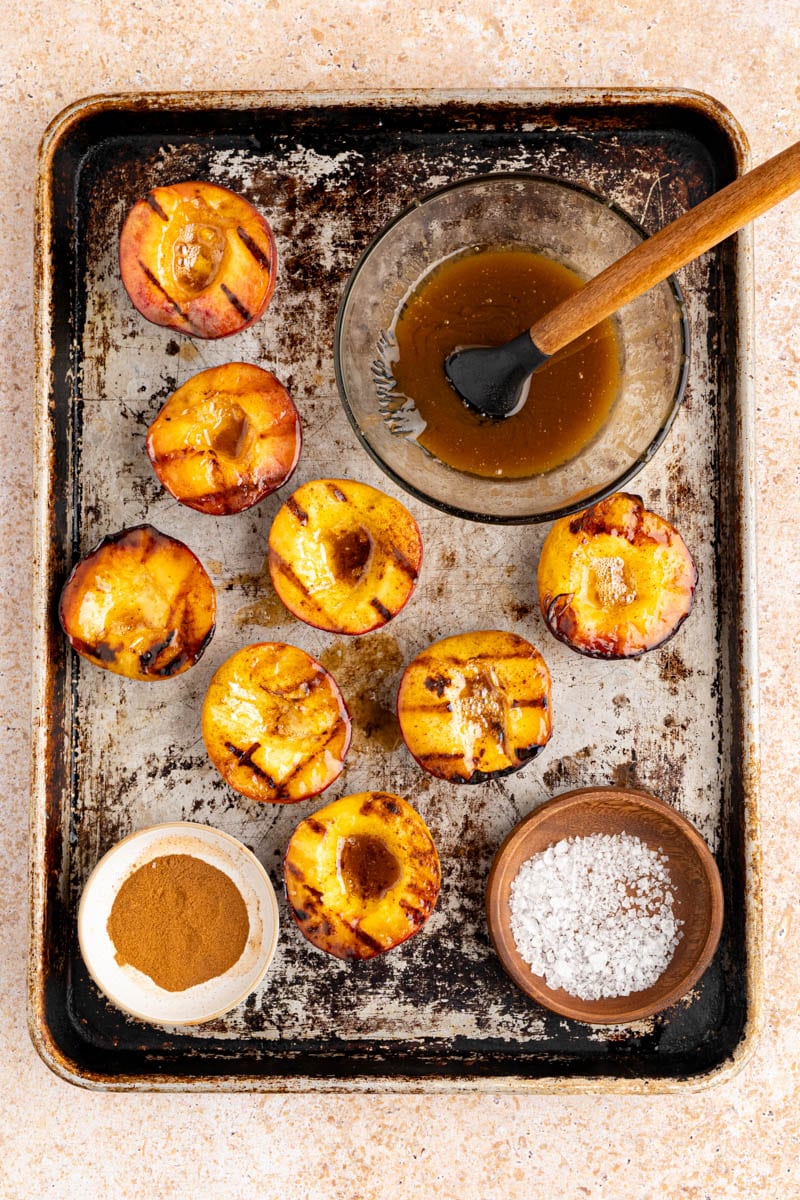 grilled peaches on a baking sheet with flaky sea salt, cinnamon and brown sugar butter on the side in bowls.