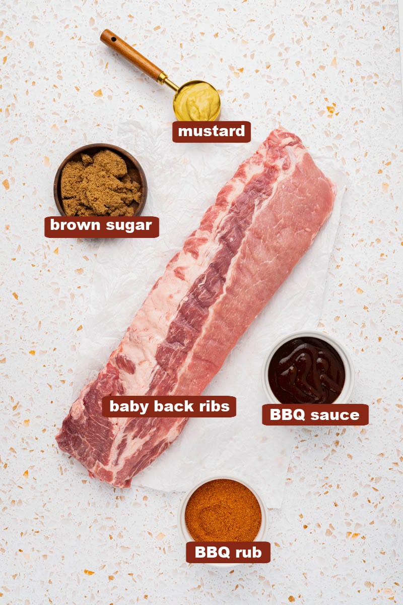 All of the ingredients for ribs on a white countertop including baby back ribs, bbq rub, mustard, brown sugar and bbq sauce. 