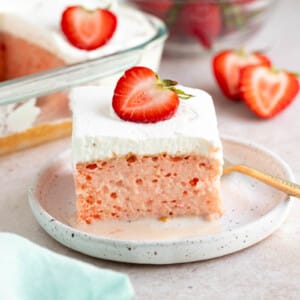 A slice of strawberry tres leches cake on a white plate for serving. Sliced strawberries and a bowl of strawberries sits to the site on a countertop.