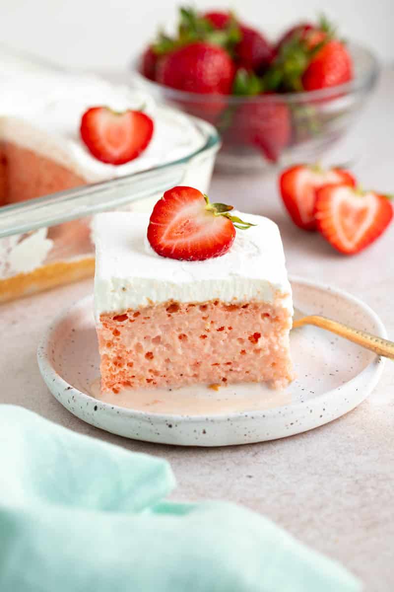 A slice of strawberry tres leches cake on a white plate for serving. Sliced strawberries and a bowl of strawberries sits to the site on a countertop.