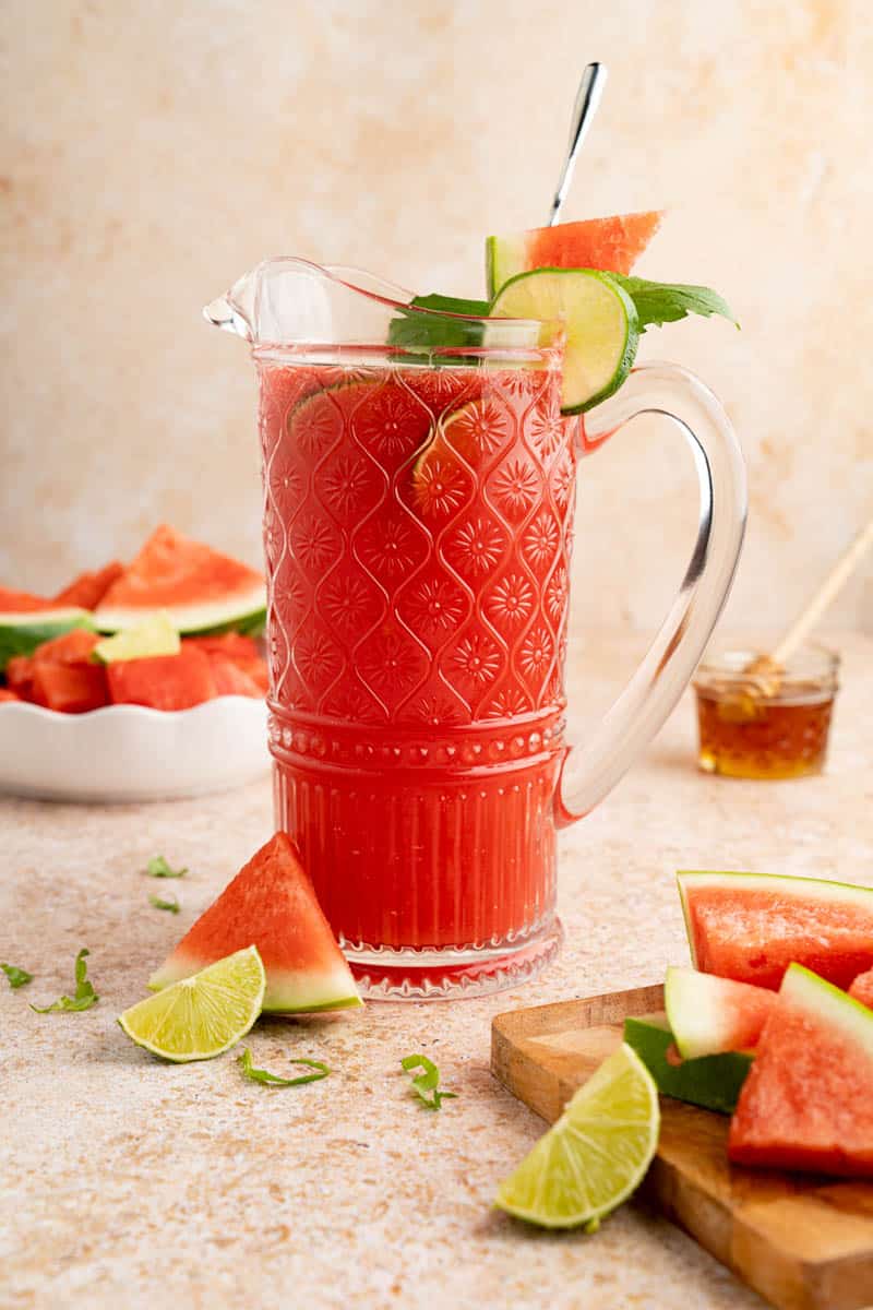 A large pitcher filled with watermelon agua fresca for serving. It's garnished with a sliced lime and fresh mint.