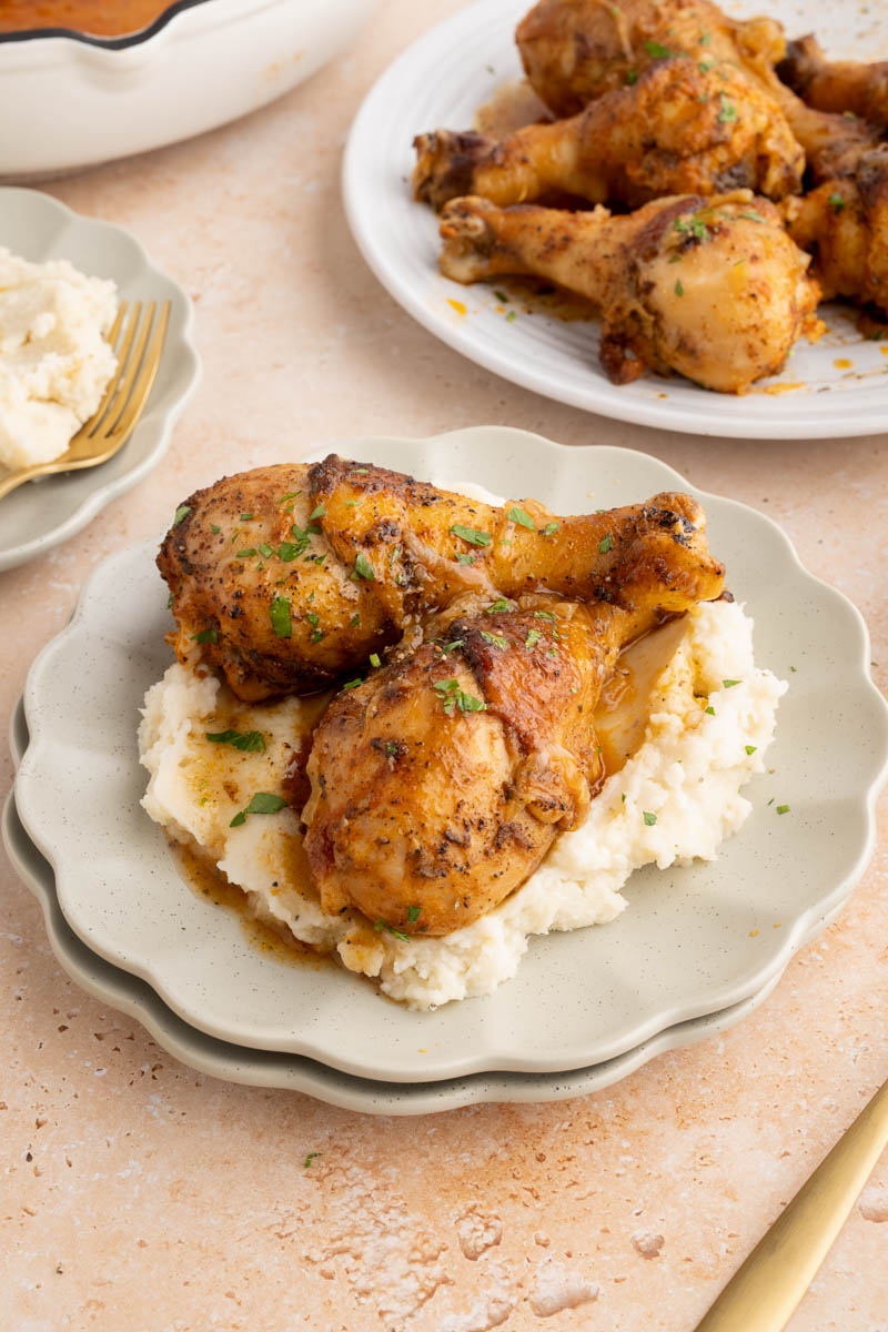 Two chicken drumsticks sit on a plate of mashed potatoes for serving. The remaining baked chicken legs sit on a serving plate on the table.