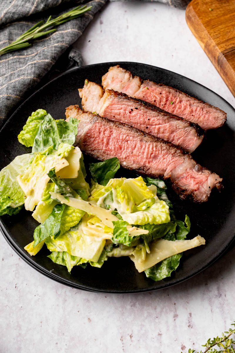 Sliced ribeye steak on a black plate with a side of caesar salad.