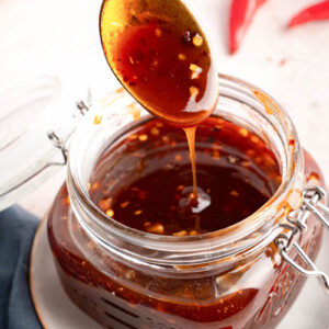 A spoon lifts a spoonful of homemade sweet chili sauce from a glass jar, The sauce drizzles off the spoon.
