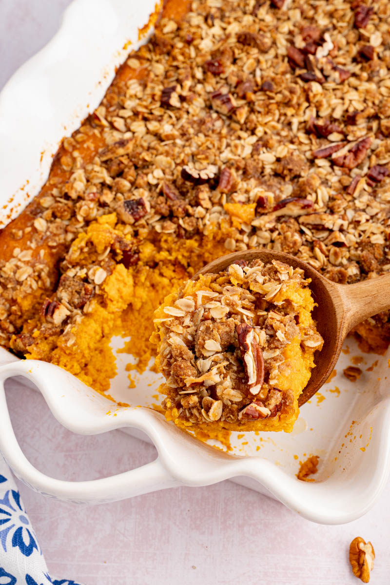 A spoon scoops out a serving of sweet potato casserole from a white rectangular baking dish.