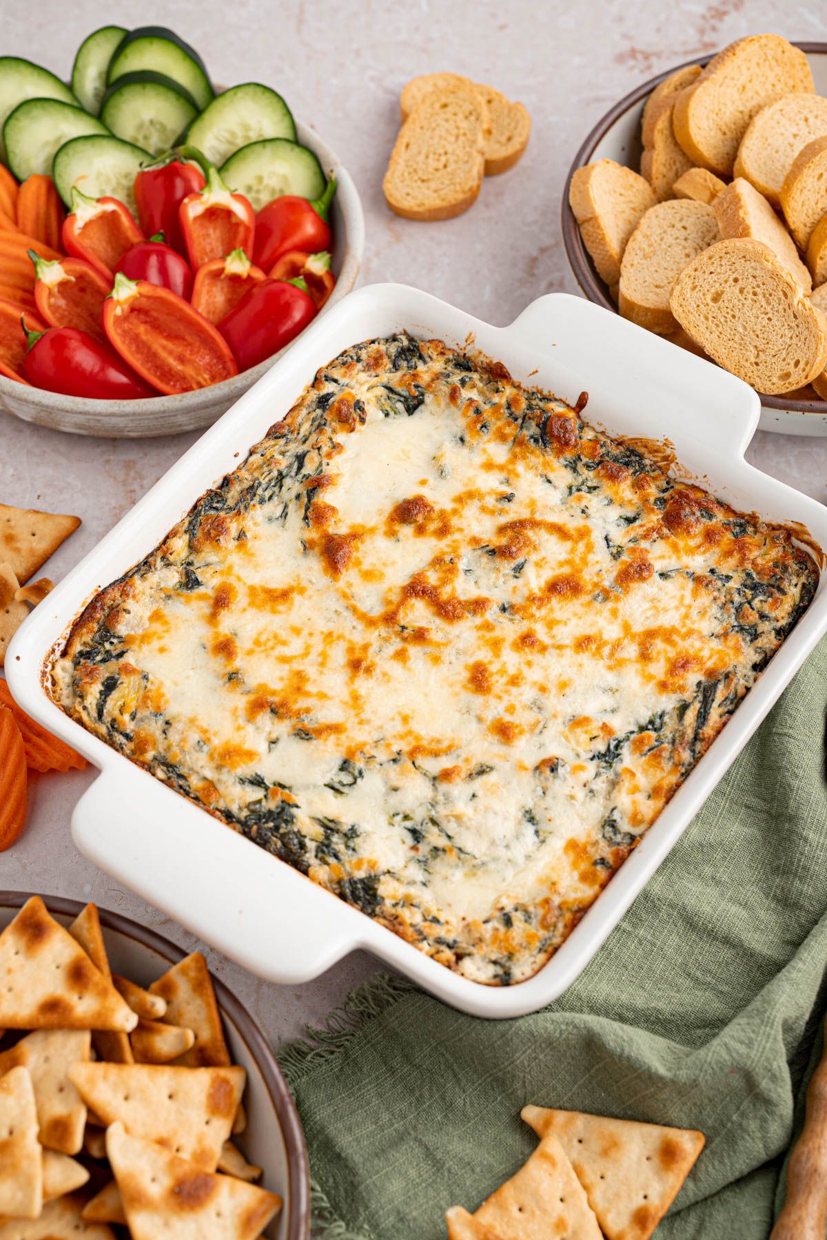 The baking dish filled with spinach and artichoke dip in a table for serving with crusty baguette and fresh sliced veggies on a plate to the side.