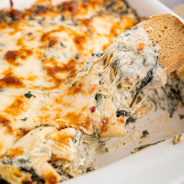 a white dish with spinach artichoke dip and a piece of toasted bread with the cheesy dip on it.