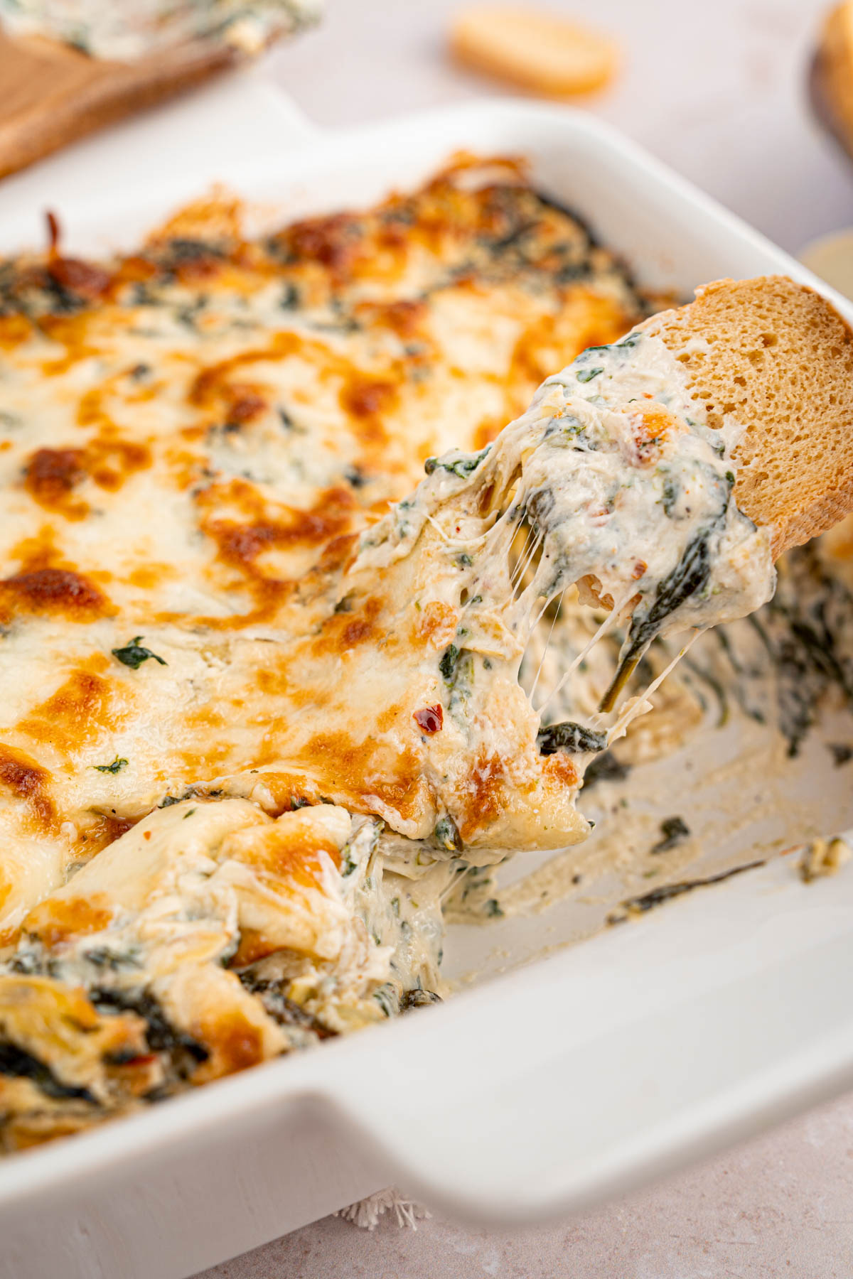 a white dish with spinach artichoke dip and a piece of toasted bread with the cheesy dip on it.