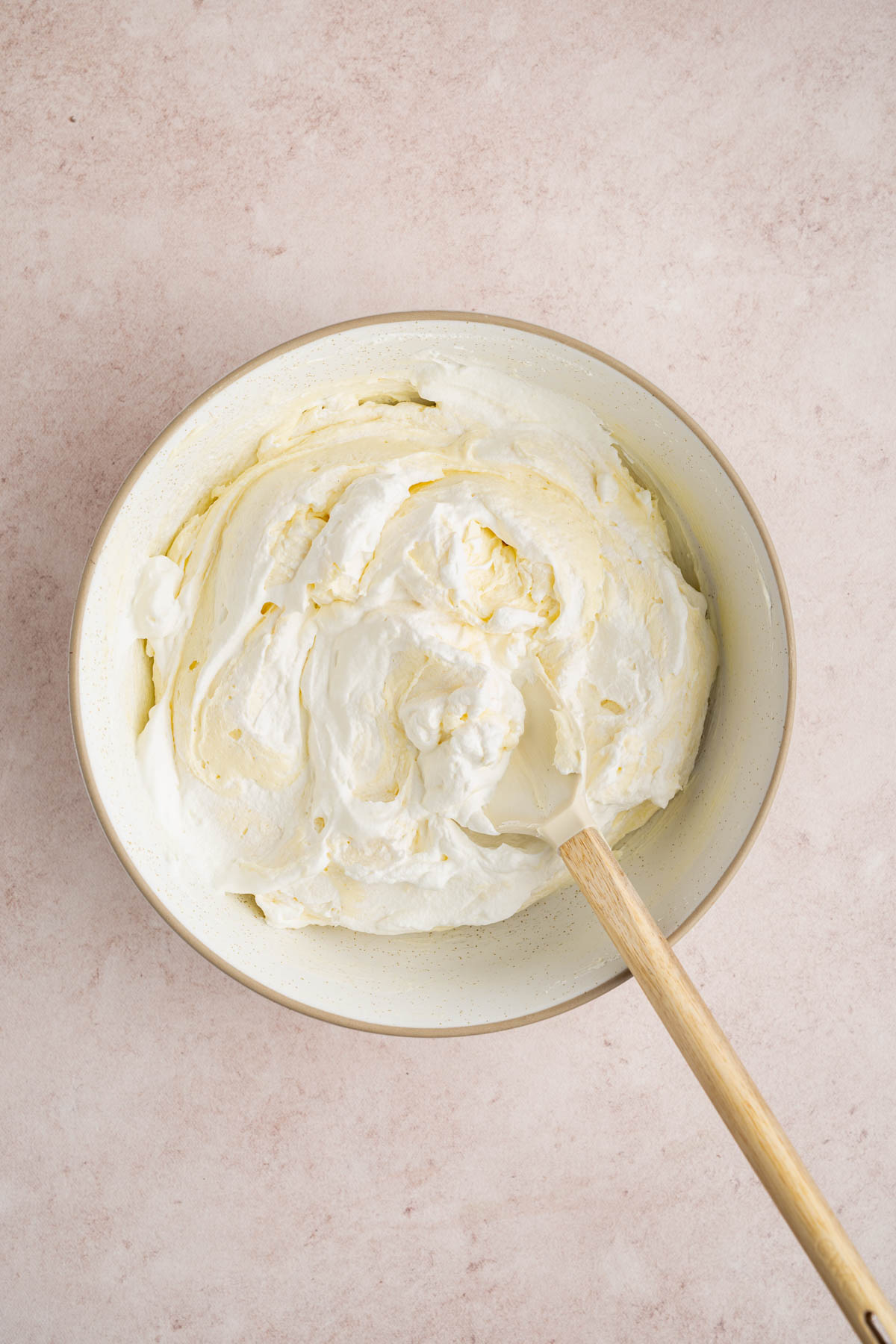 The whip cream folded into the cream cheese mixture in the white bowl on the countertop. A wooden spoon sits in the bowl.