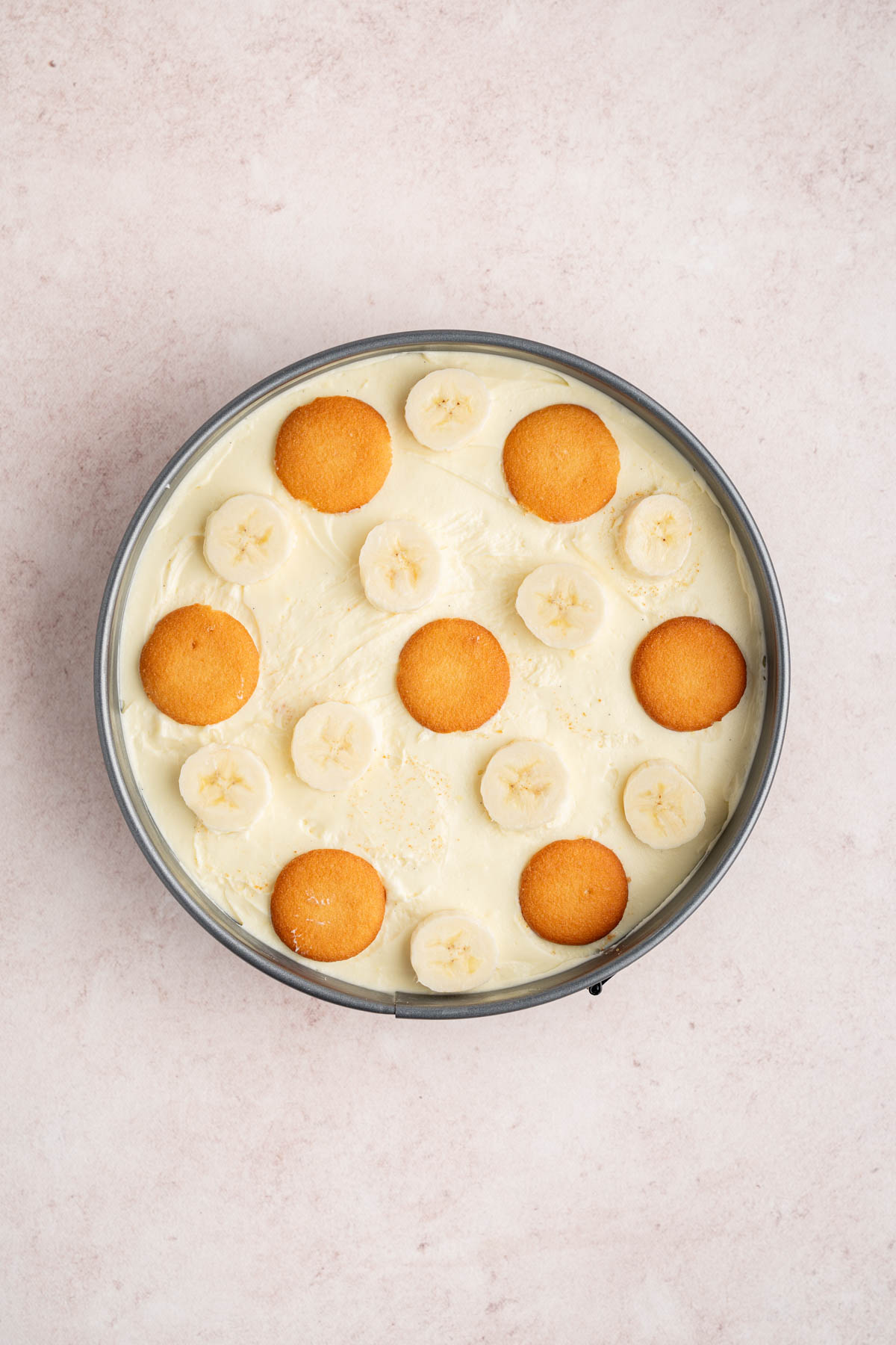 Whole nilla wafers and banana slices over the top of the cheesecake filling in the pan on a countertop.