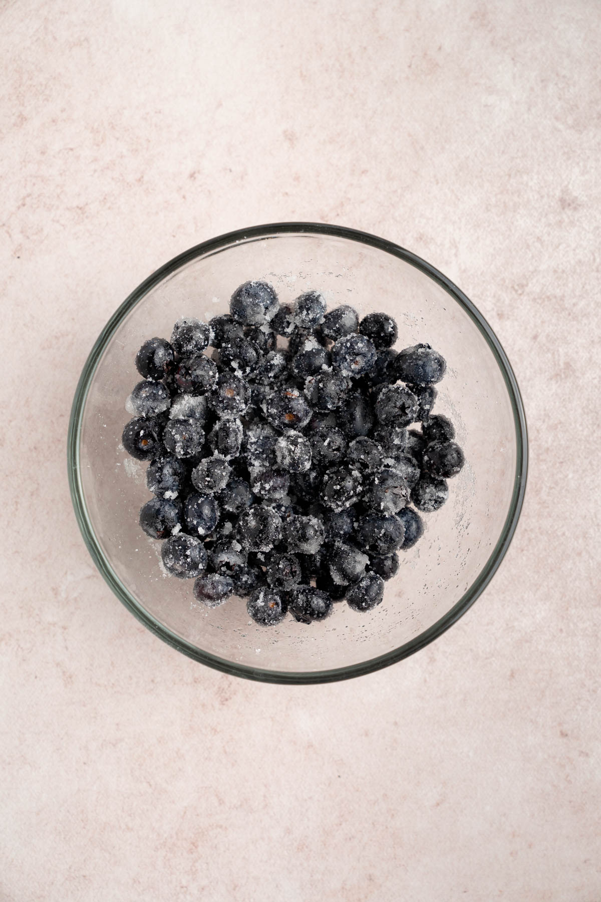 Blueberries and sugar in a clear glass bowl on a countertop after mixing together.