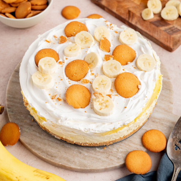 No Bake Banana Pudding Cheesecake on a cake serving plate sitting on a countertop. Bananas and extra vanilla wafers are in a bowl on the side.