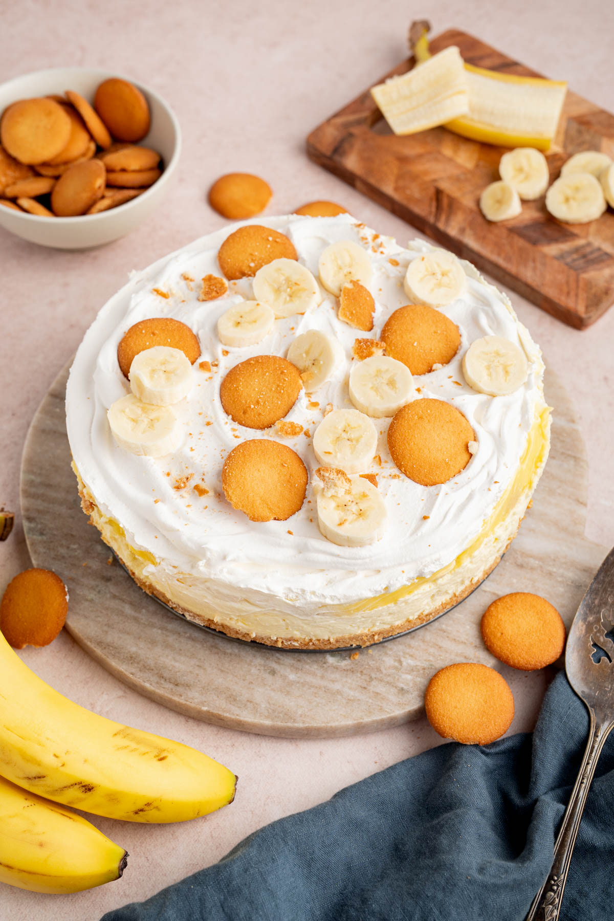 No Bake Banana Pudding Cheesecake on a cake serving plate sitting on a countertop. Bananas and extra vanilla wafers are in a bowl on the side.