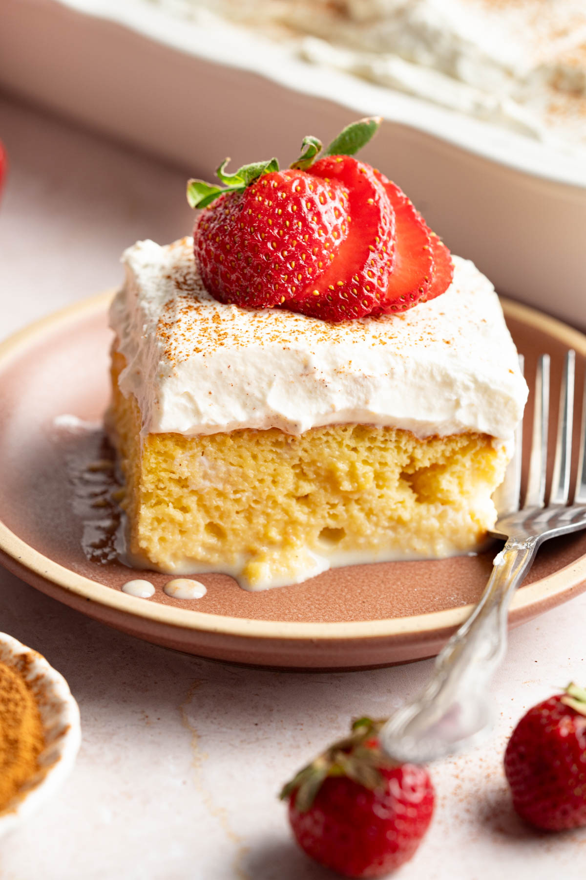 A slice of tres leches cake with a sliced strawberry on top on a plate with a fork for serving.