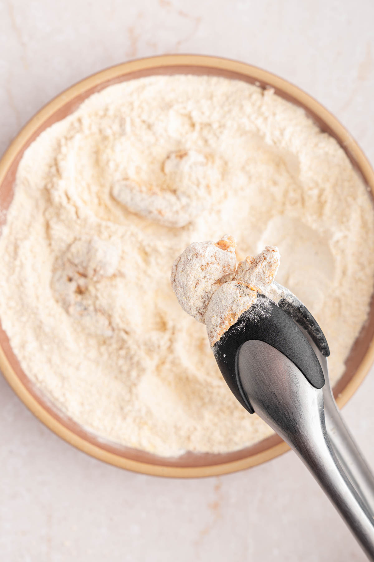 Tongs hold a piece of shrimp that is dredged in the flour mixture before frying.