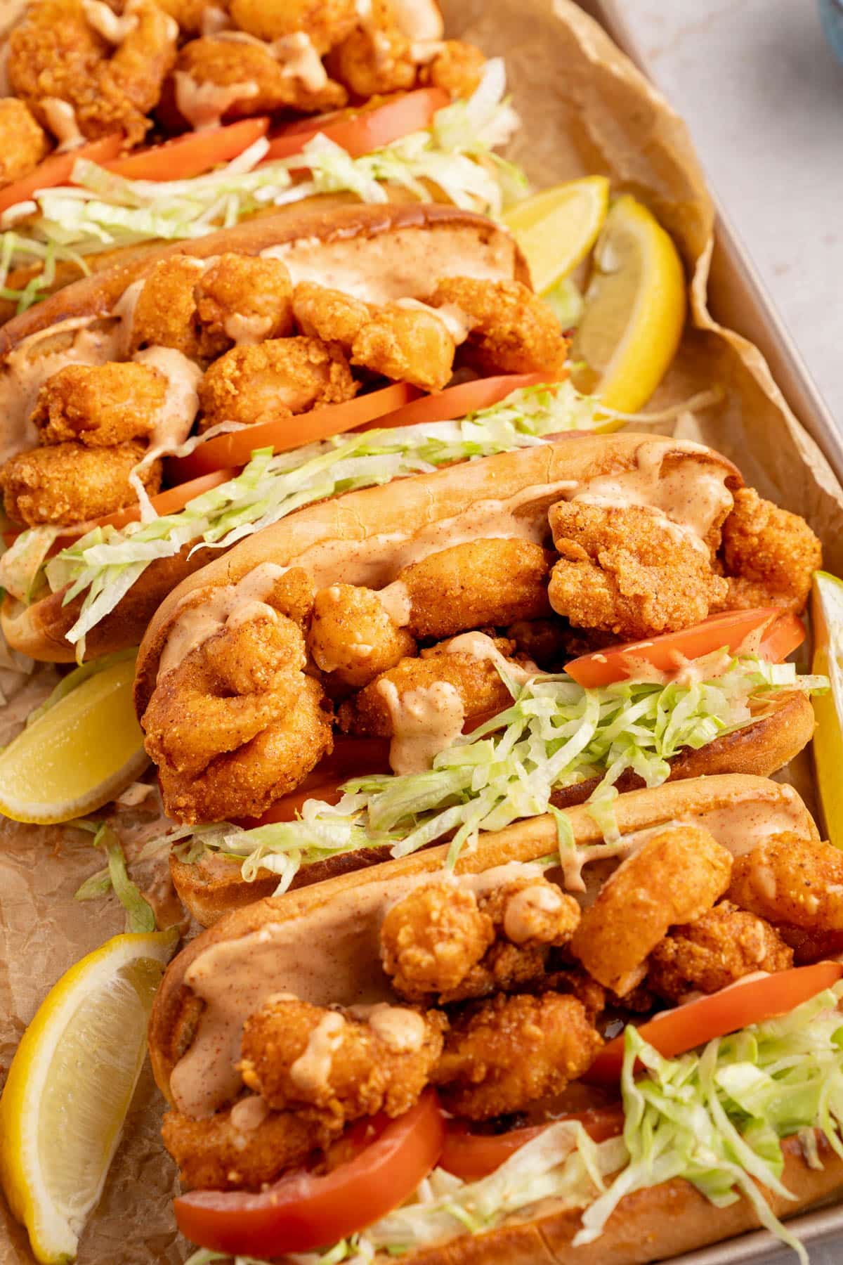 Four shrimp po boys on a baking sheet after being assembled with lemon wedges on the side.
