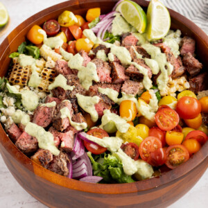 The steak salad with cilantro-lime avocado crema drizzled on top in a wooden bowl on a white countertop.