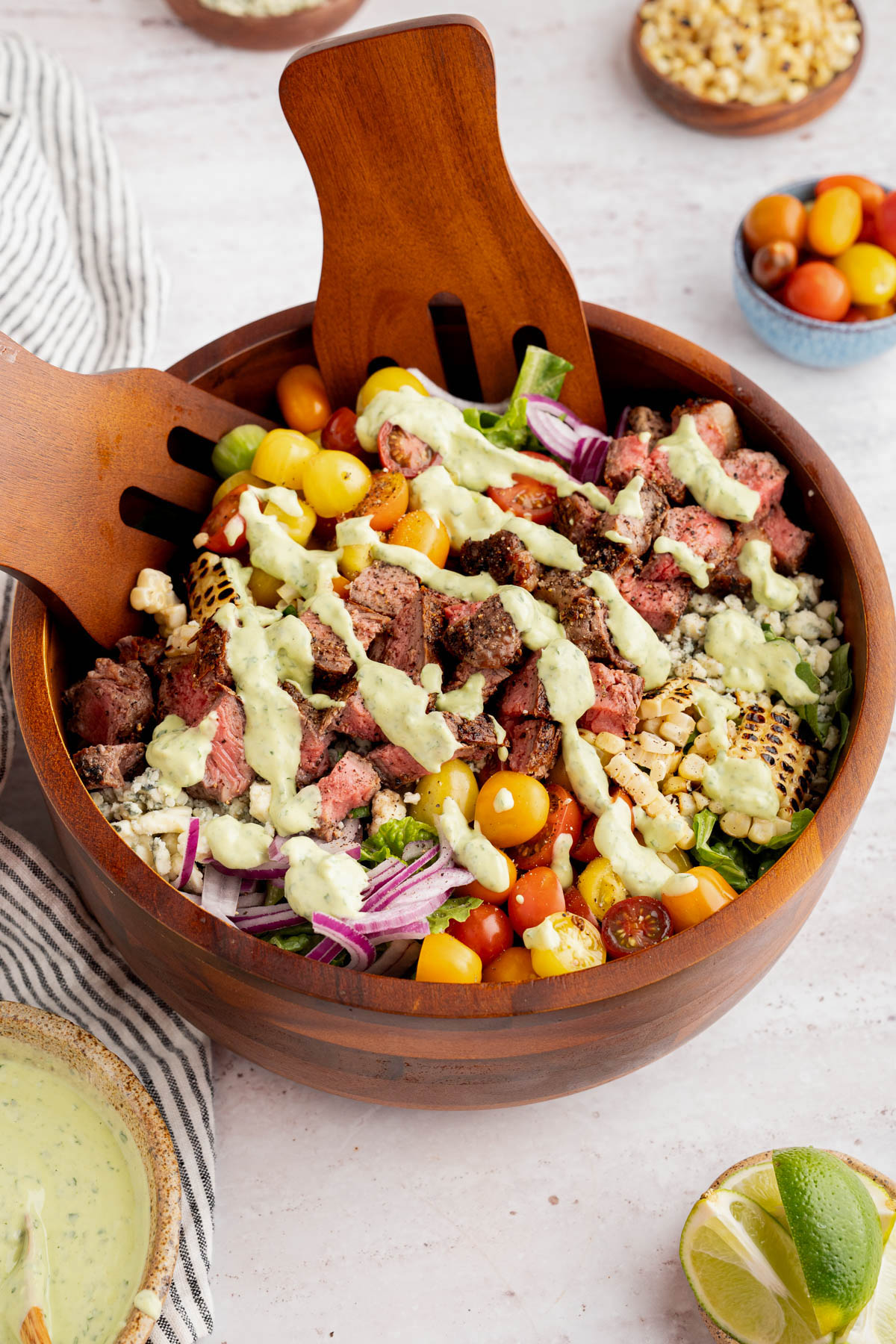 The steak salad with the avocado-lime cilantro crema drizzled on top in a bowl on a countertop. Two serving forks sit inside the bowl.