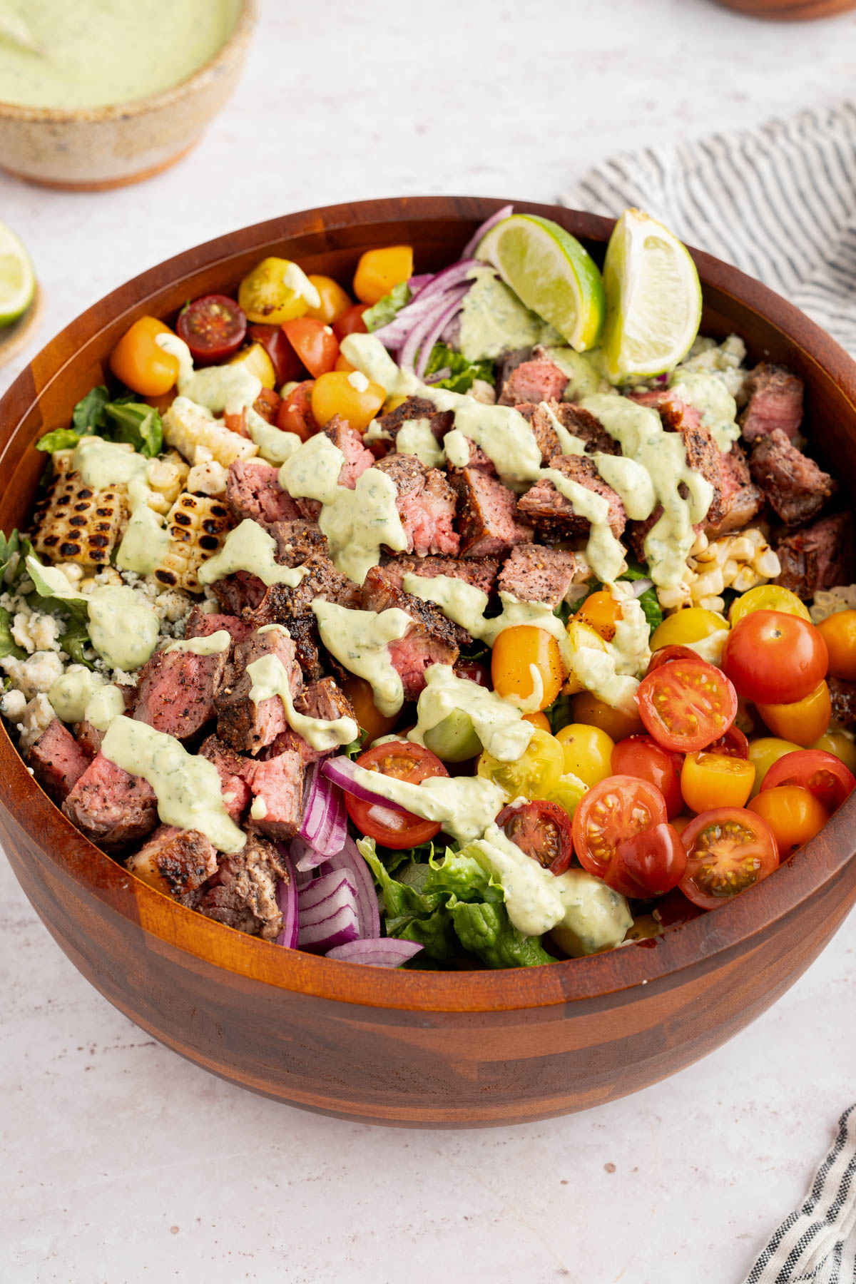 The steak salad with cilantro-lime avocado crema drizzled on top in a wooden bowl on a white countertop.