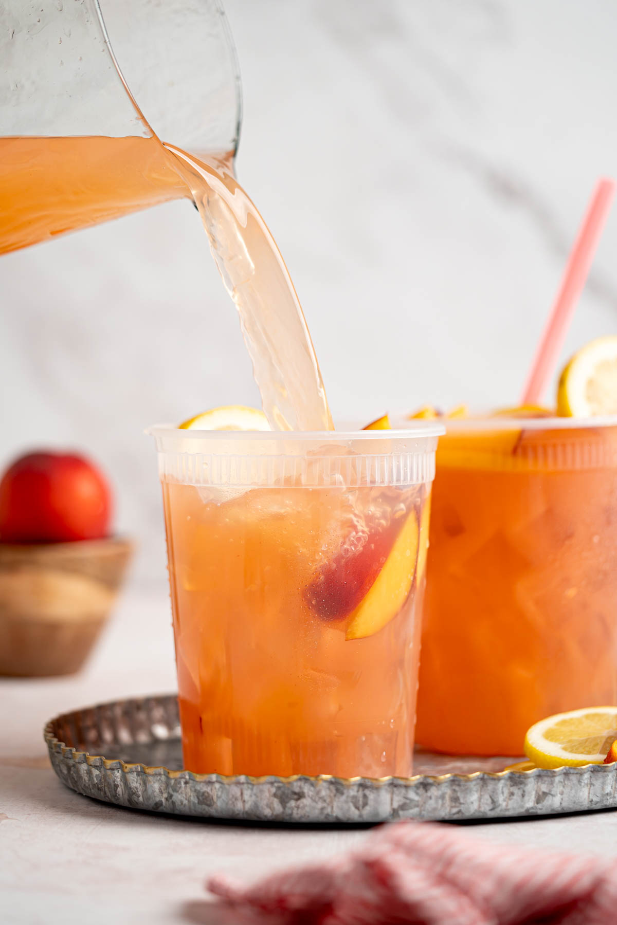 A pitcher pours the peach lemonade into a party cup filled with ice, sliced peaches, and lemon wheels.