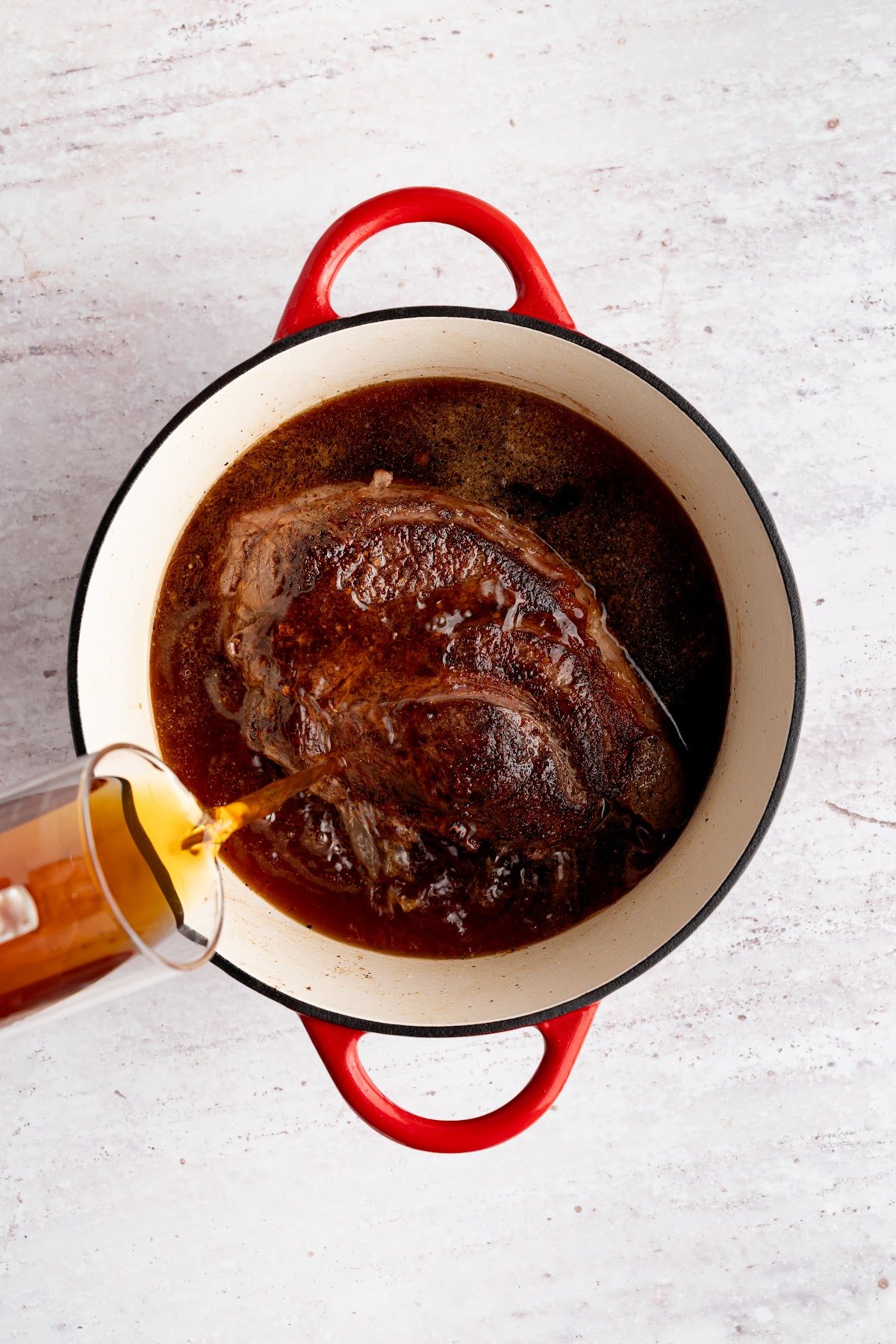 Beef broth being poured into the Dutch oven around the seared chuck roast to braise it and become the au jus