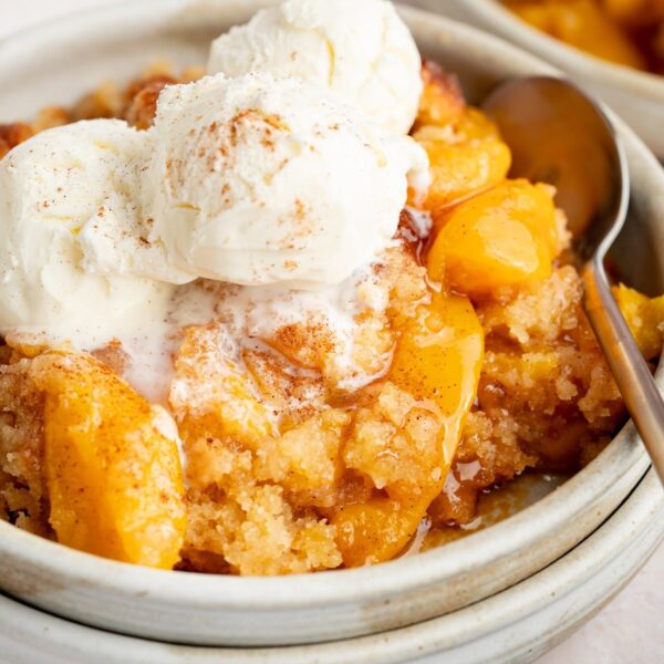 Biscuit top peach cobbler served warm in a bowl with two scoops of vanilla ice cream melting over the peaches