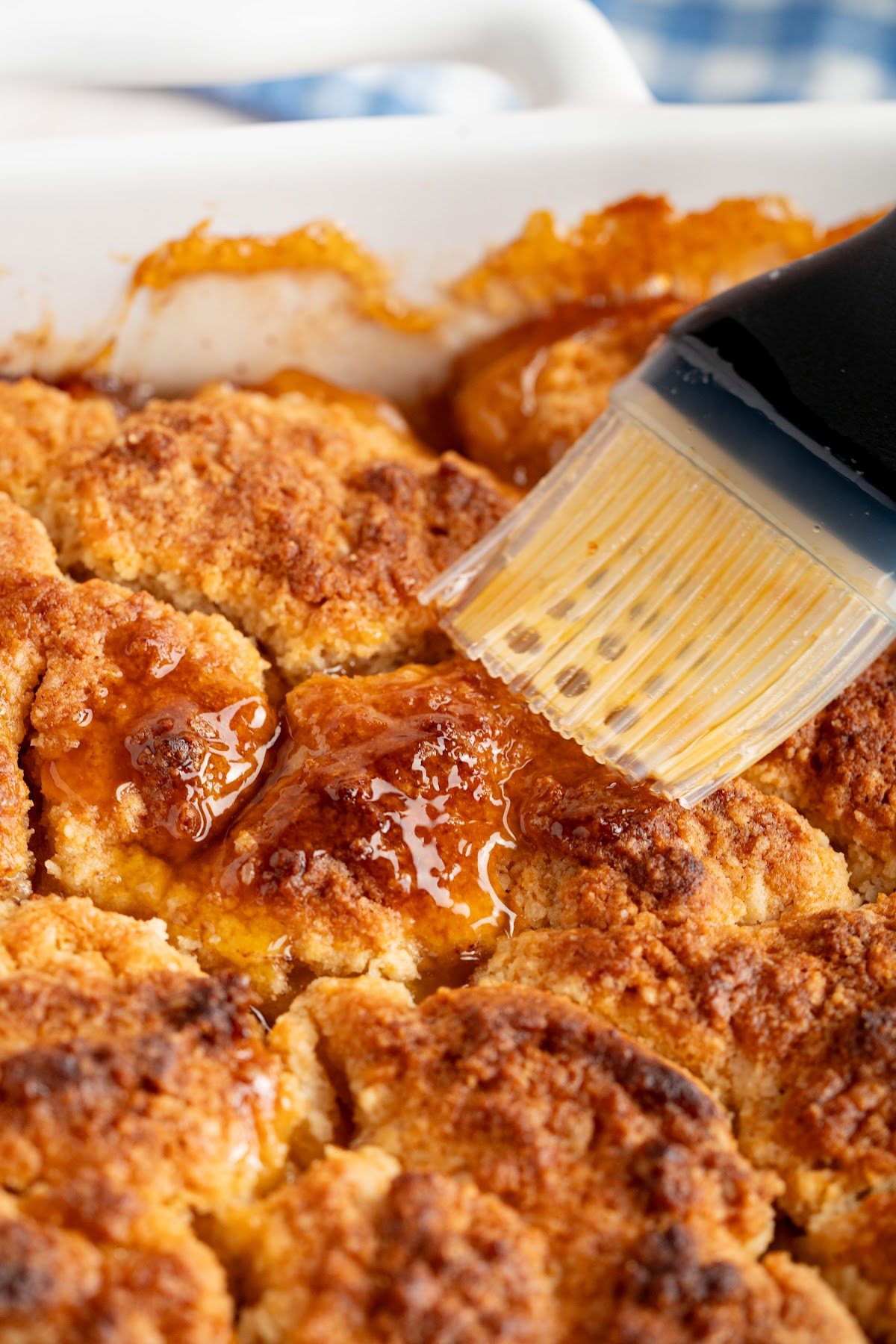 A pastry brush spreading honey butter over the golden brown biscuit top of a peach cobbler