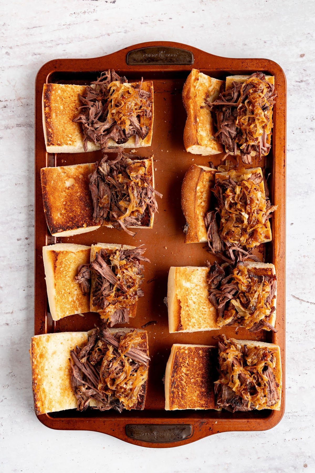 Split baguettes on a sheet pan piled with shredded beef and caramelized onions
