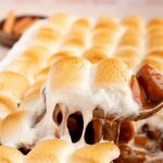 A spoon lifting a scoop of candied yams with toasted marshmallows stretching from the pan