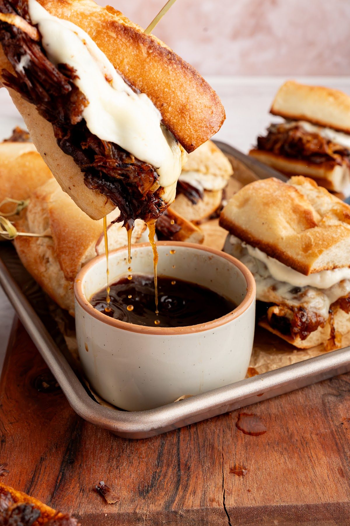 A French dip sandwich being dipped into a bowl of hot au jus on a sheet pan