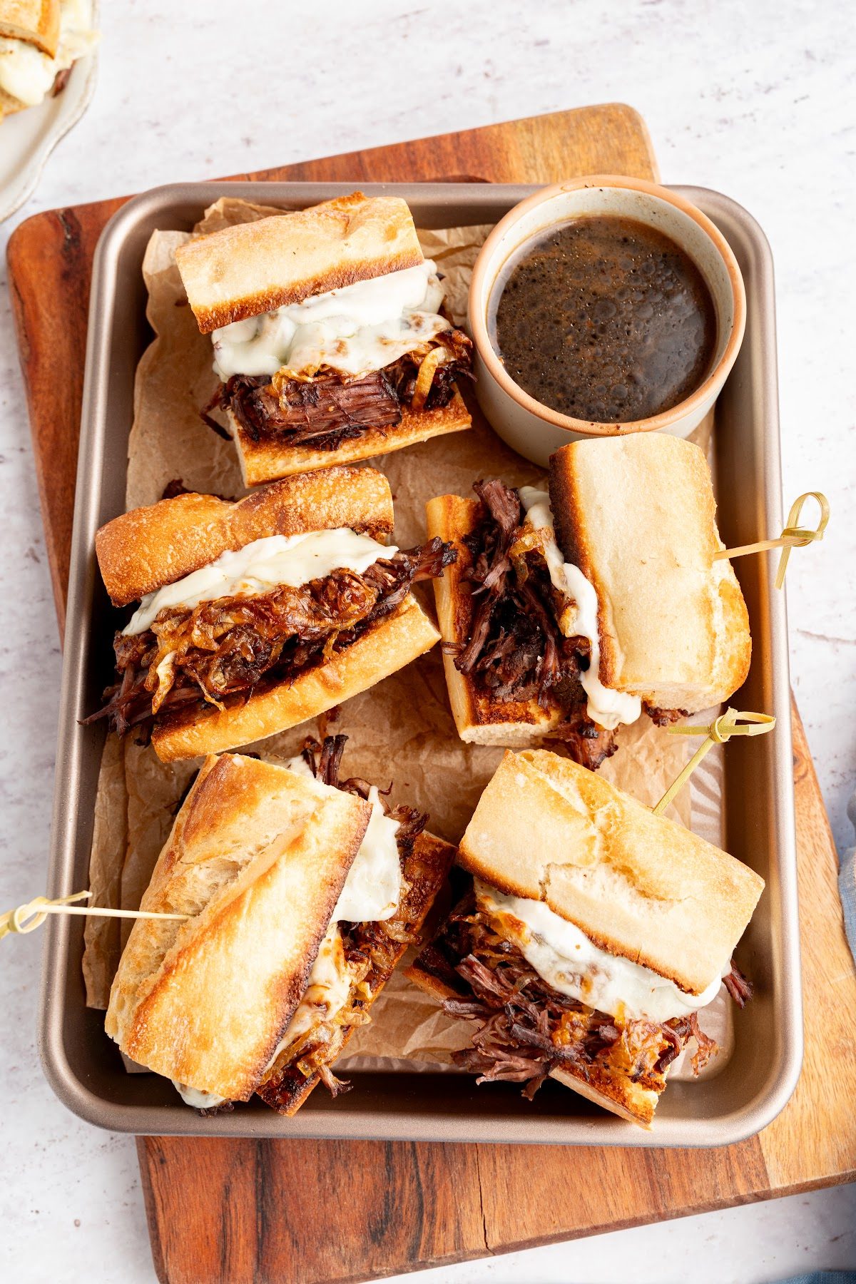 Four French dip sandwiches cut and skewered on a sheet pan with a bowl of au jus in the middle