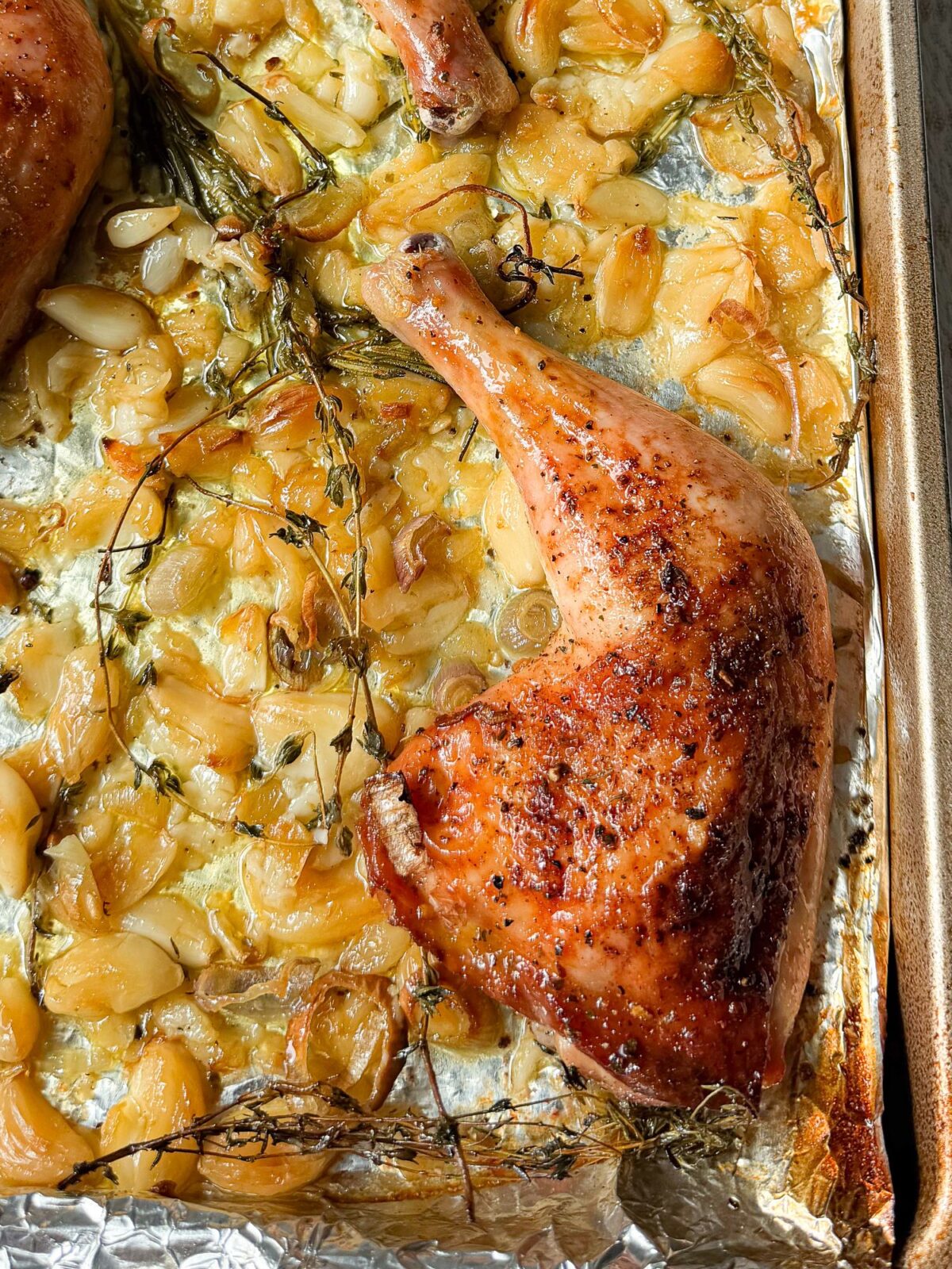 Crispy golden garlic confit chicken leg quarter on a sheet pan with roasted garlic cloves and fresh thyme