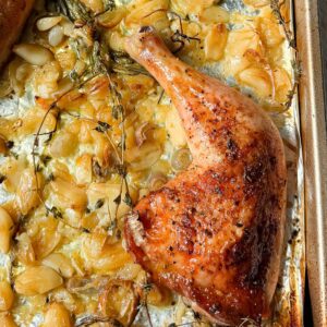 Crispy golden garlic confit chicken leg quarter on a sheet pan with roasted garlic cloves and fresh thyme