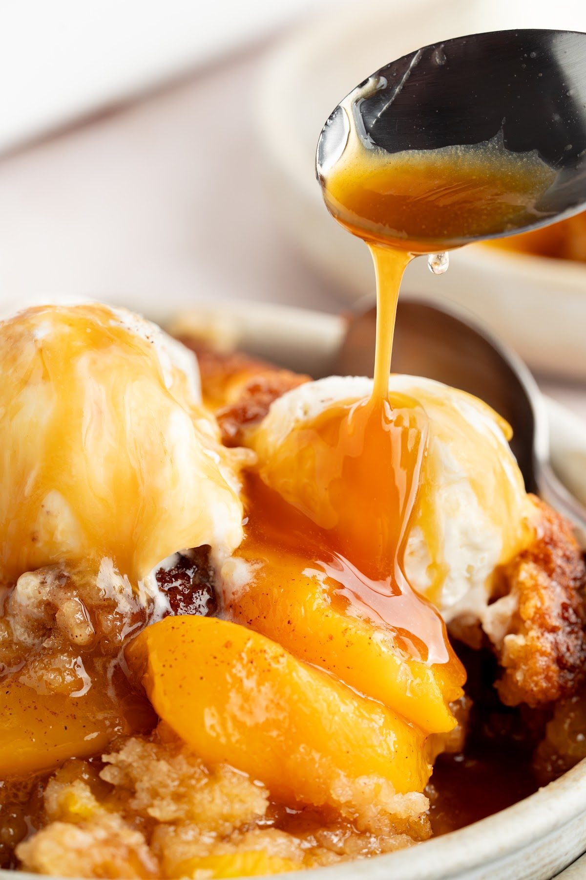 Warm peach syrup being spooned over vanilla ice cream on a serving of biscuit top peach cobbler