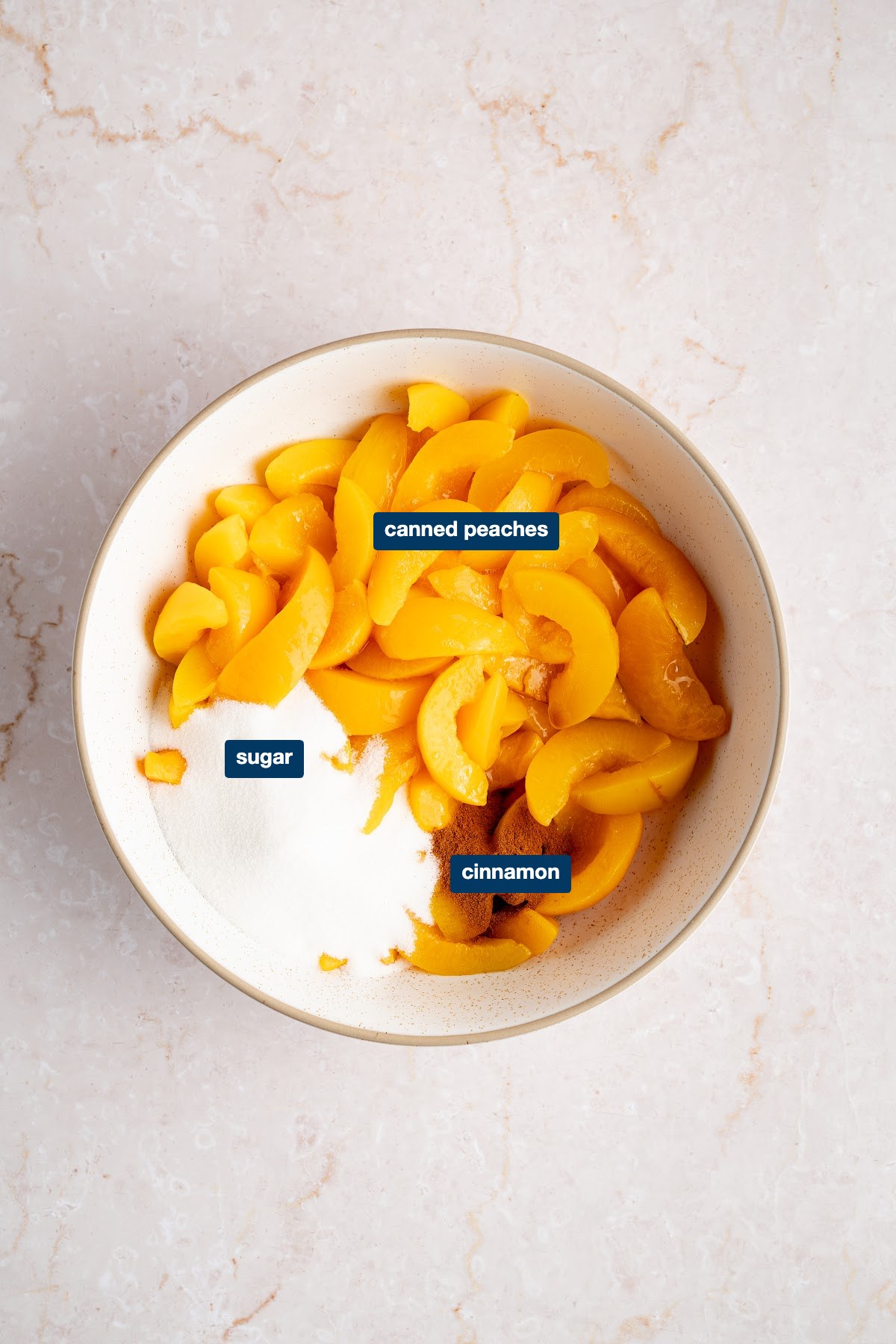 Labeled ingredients for peach cobbler filling: canned sliced peaches, granulated sugar and ground cinnamon in a bowl