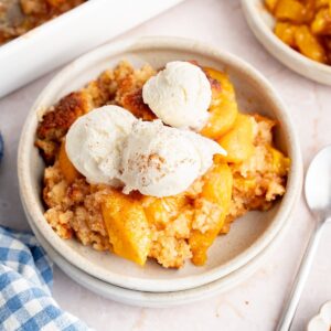 A bowl of warm biscuit top peach cobbler with a scoop of vanilla ice cream next to the baking dish