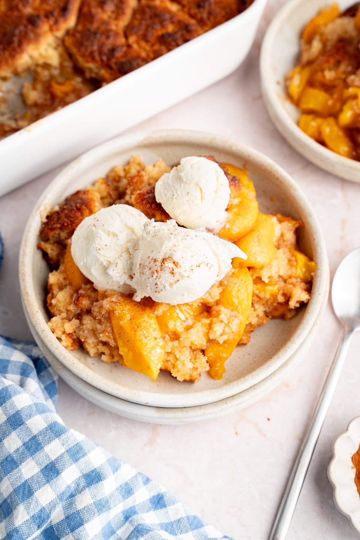 A bowl of warm biscuit top peach cobbler with a scoop of vanilla ice cream next to the baking dish