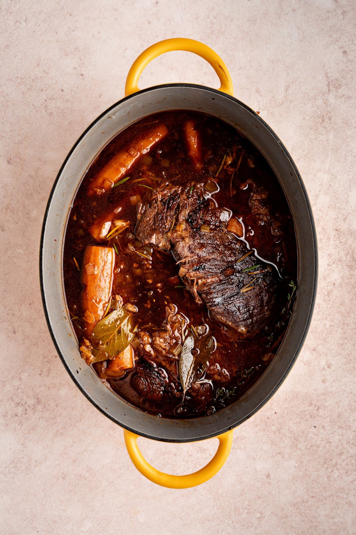 A seared chuck roast back in the Dutch oven with a whole garlic bulb, herbs and halved carrots laid on top of the red wine braising liquid